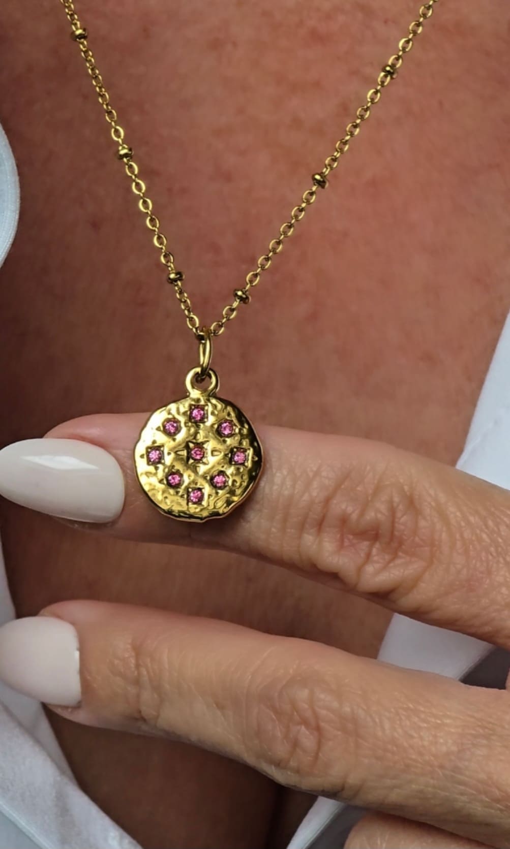 Collier doré porté au cou, médaillon martelé orné d’un strass rose sur chaîne perlée, création artisanale Zateka