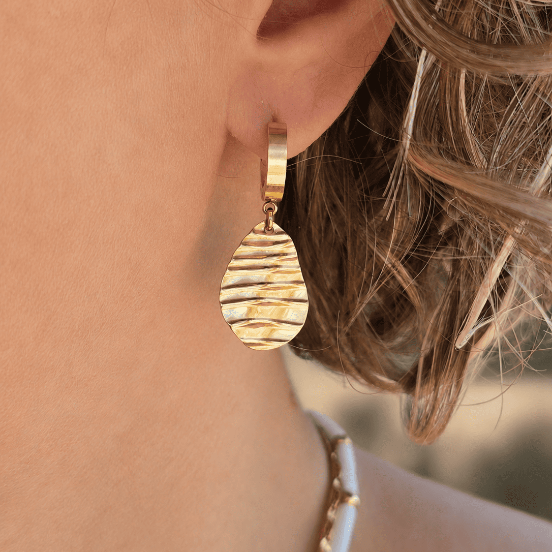 Boucles d'oreilles créoles dorées portées avec pendentif effet ondulé.
