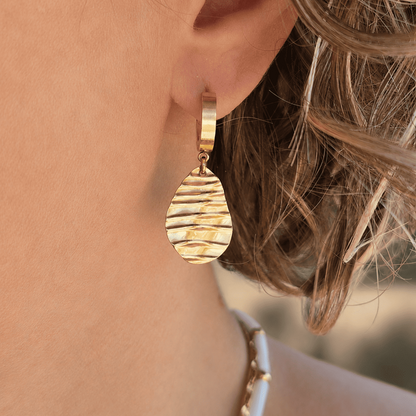 Boucles d'oreilles créoles dorées portées avec pendentif effet ondulé.
